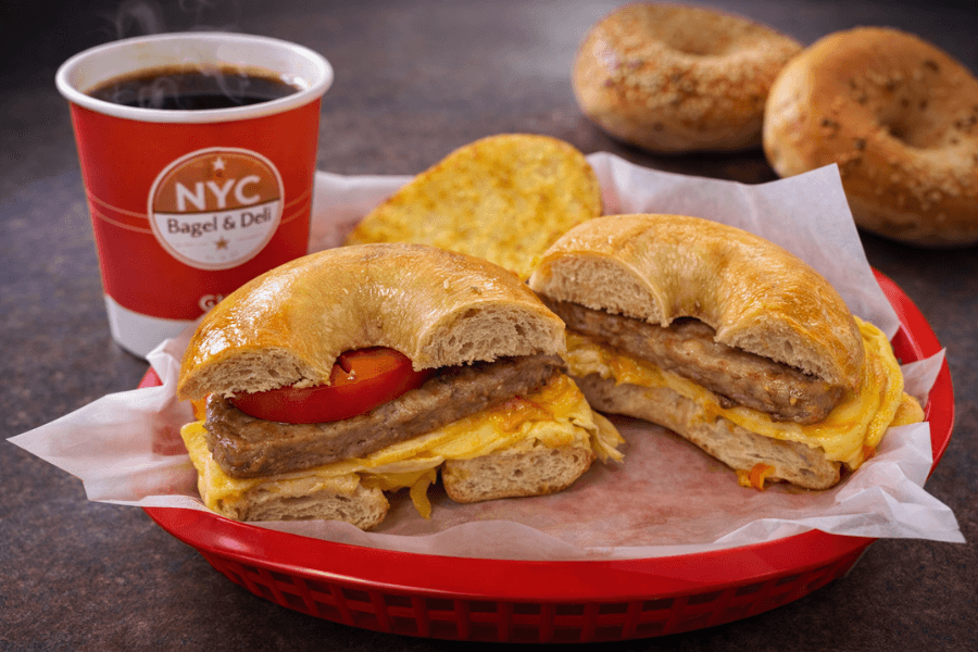 Bagel sandwich with sausage, egg, cheese, and tomato in a red basket, with coffee and bagels nearby.