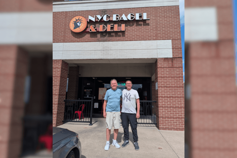 Two men stand smiling outside the entrance of a NYC Bagel & Deli storefront.