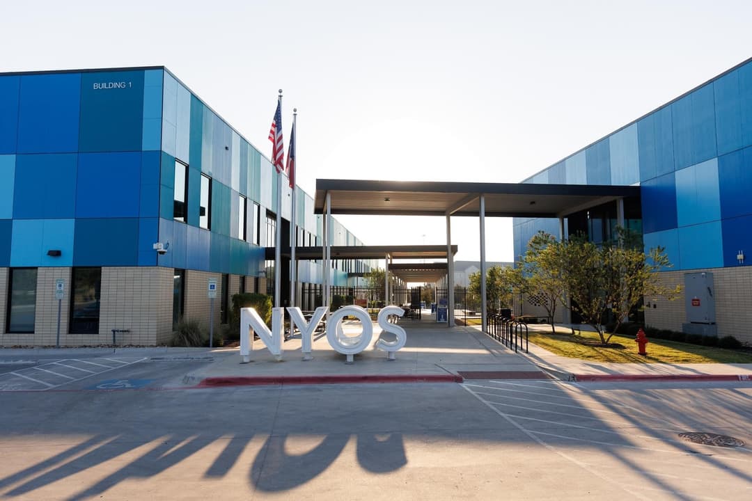 Large white "NYOS" letters outside a blue-paneled school building with US and Texas flags.