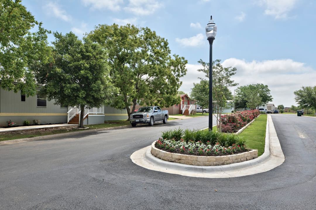 Landscaped median with flowers and lamppost on residential street, pickup truck parked near mobile homes under trees.