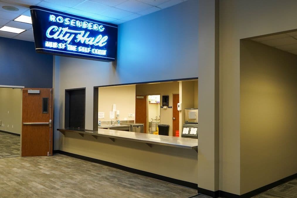 Neon "Rosenberg City Hall, Hub of the Gulf Coast" sign above a service counter and kitchen window.