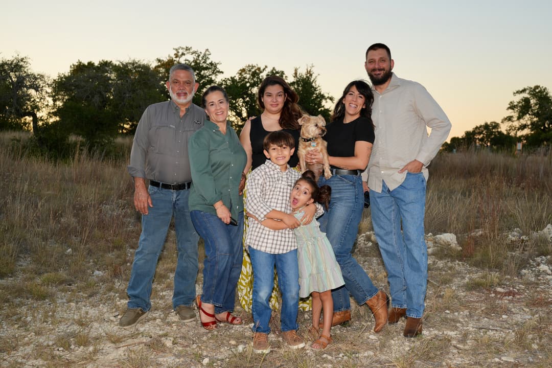 A family of seven with two children and a small dog pose together outdoors in a grassy field at dusk.