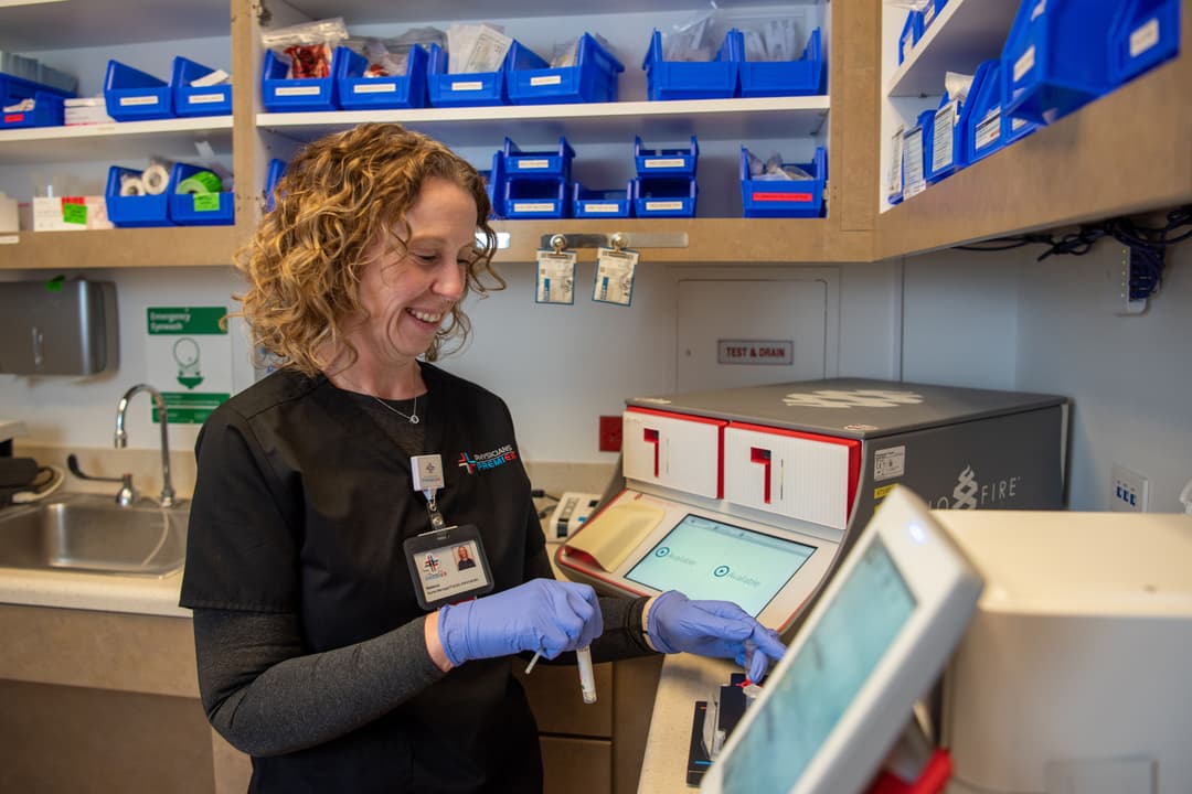 Smiling medical worker in gloves uses a lab testing machine in a clinic supply room with blue bins on shelves.