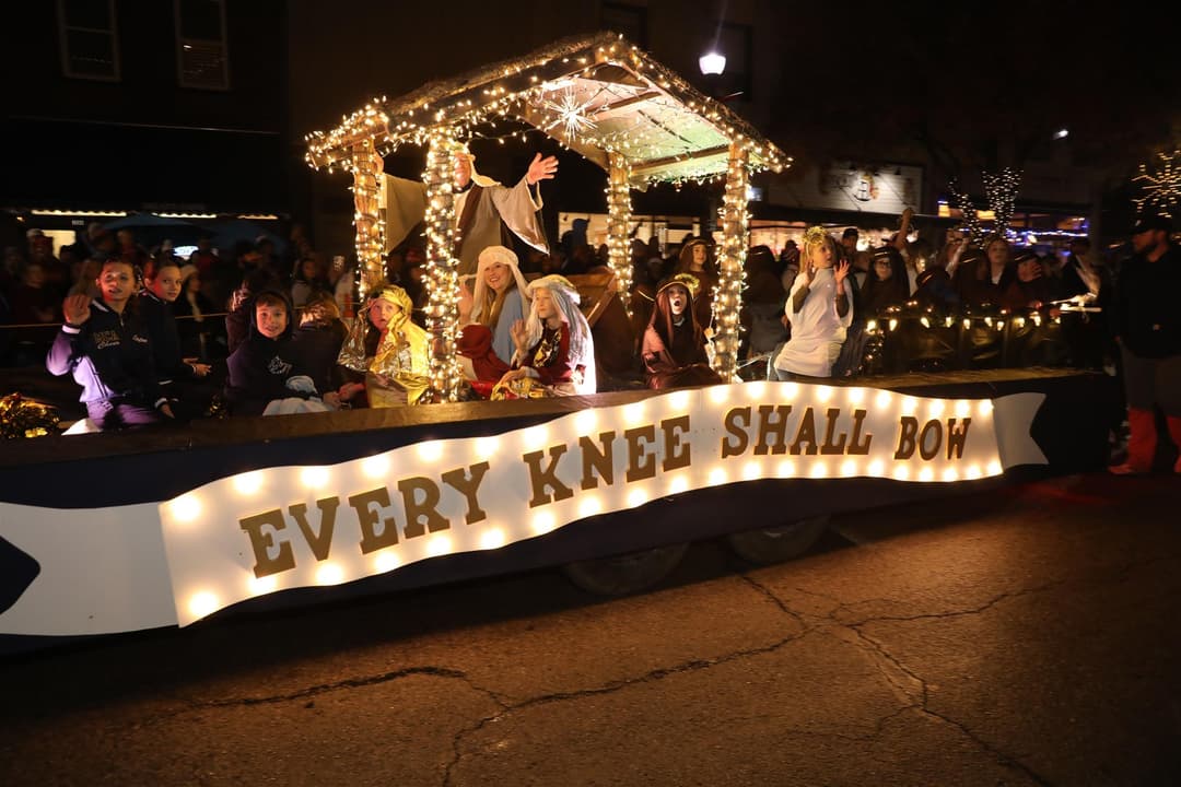 Lit-up nativity parade float with costumed children and "Every Knee Shall Bow" sign at a nighttime Christmas parade.