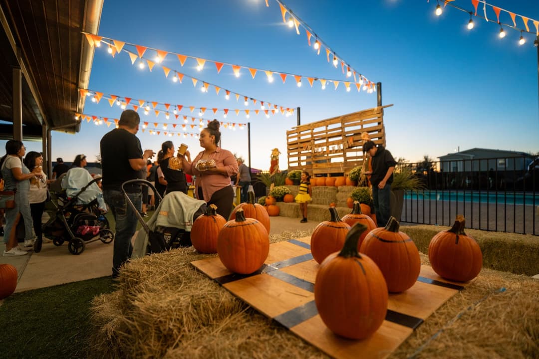 Outdoor fall festival at dusk with pumpkins on hay bales, string lights, and families gathered near a pool.