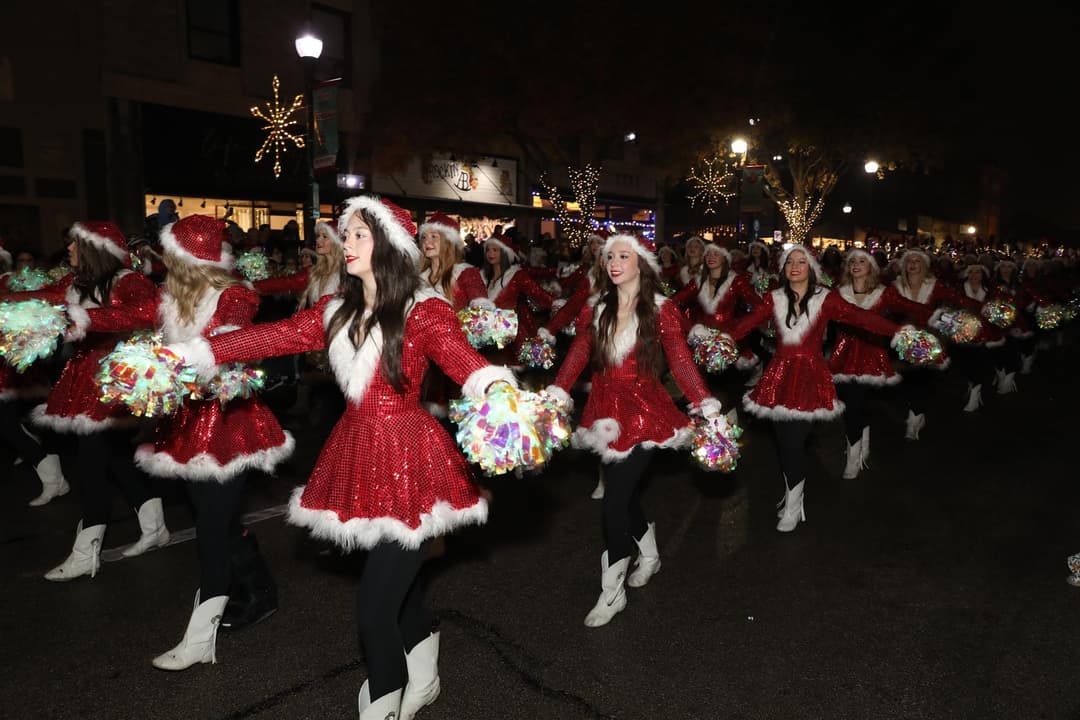 Dance team in red Santa dresses with pom-poms perform in a nighttime holiday parade downtown.