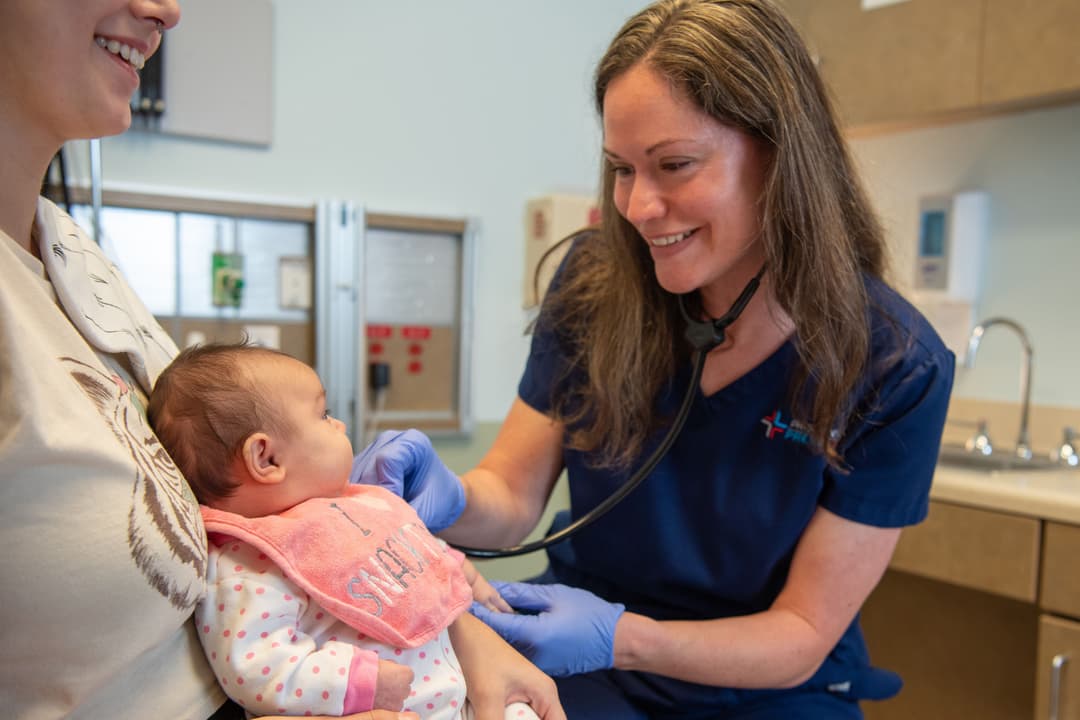 Smiling nurse in blue scrubs uses a stethoscope to examine a baby held by a parent in an exam room.