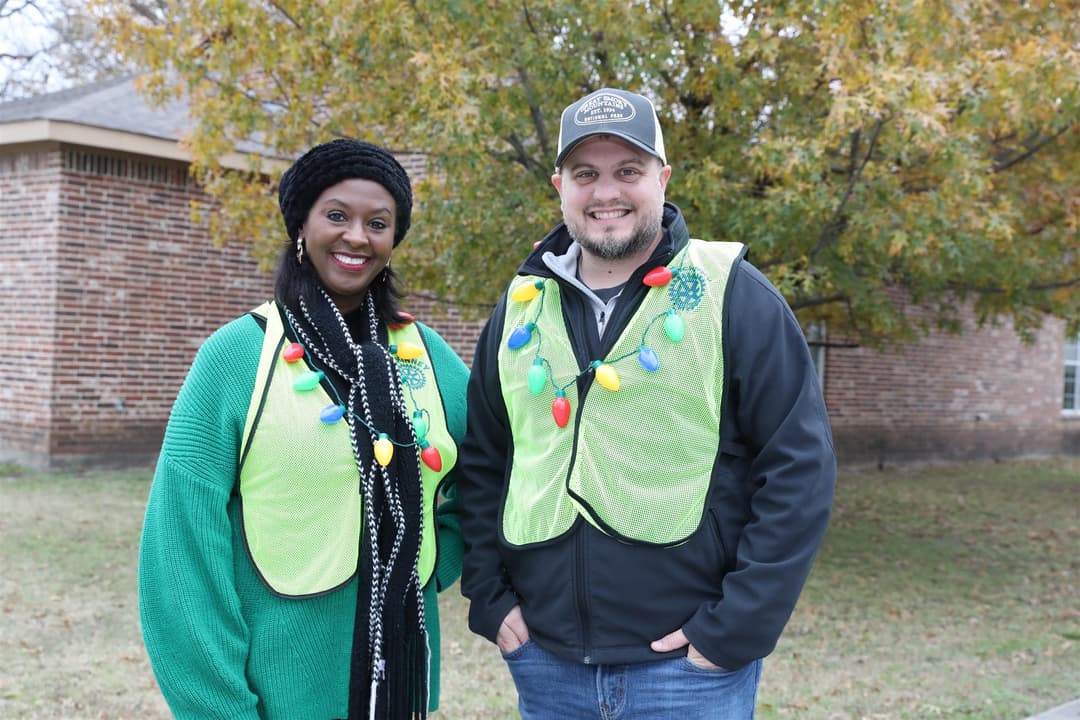 Two people in yellow safety vests with Christmas light necklaces smile outdoors by a brick building.