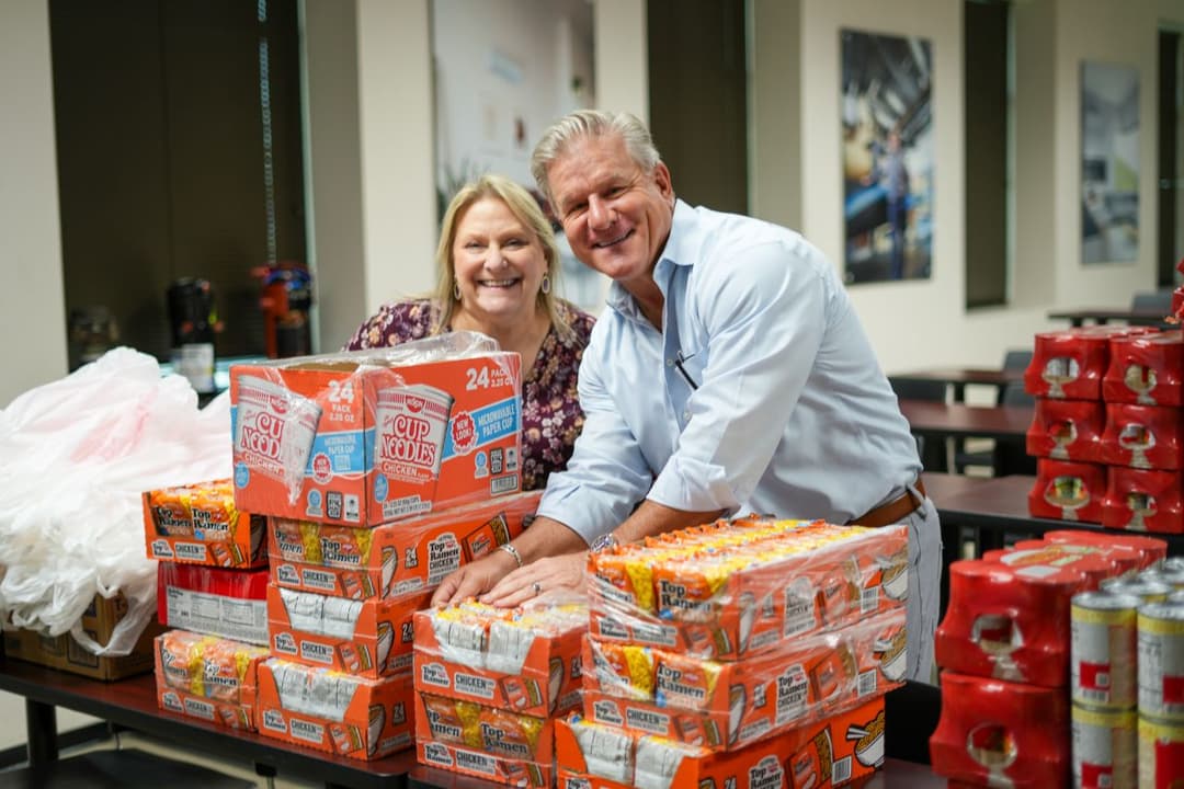 Smiling man and woman stand behind a table stacked with packaged ramen noodles and cup noodles for donation.