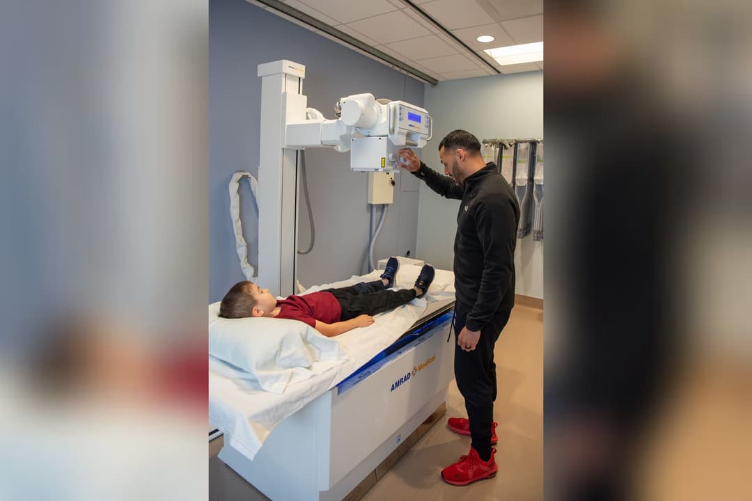 Technician adjusts an X-ray machine over a boy lying on an exam table in a medical imaging room.