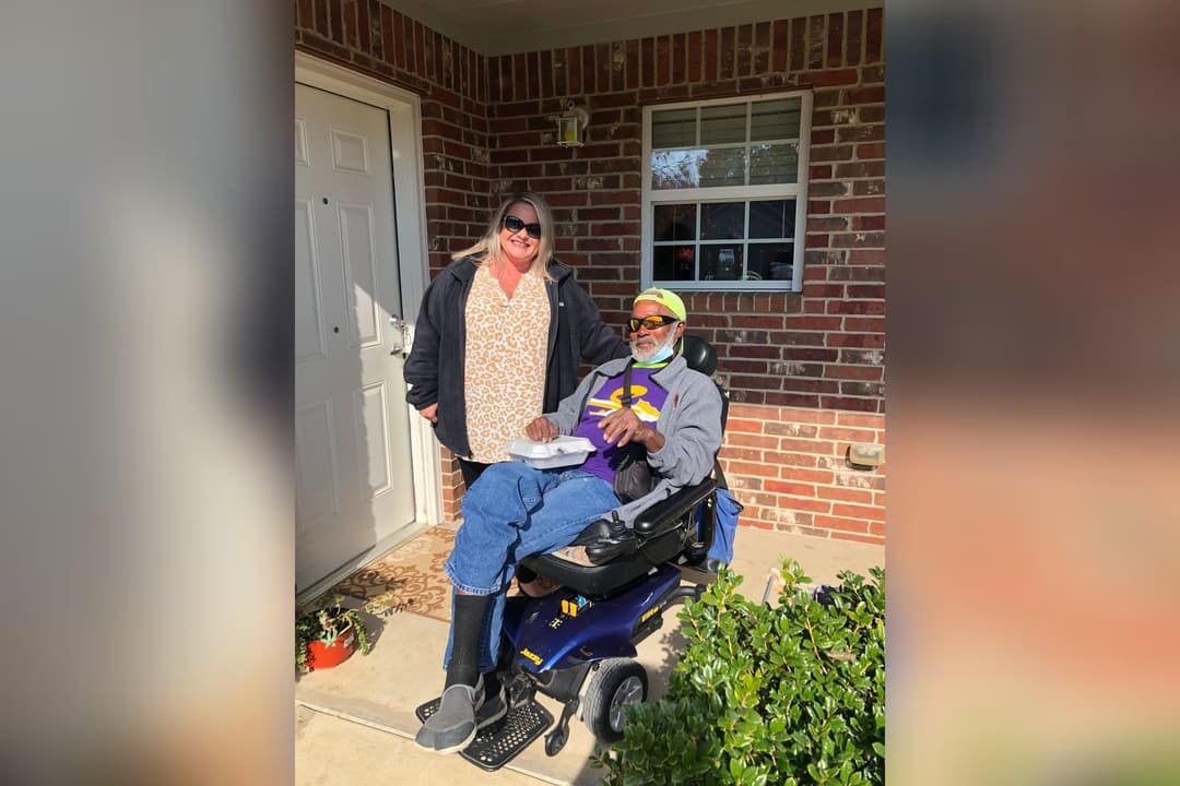Woman stands beside a man in a power wheelchair holding a meal container, on a brick home's front porch.