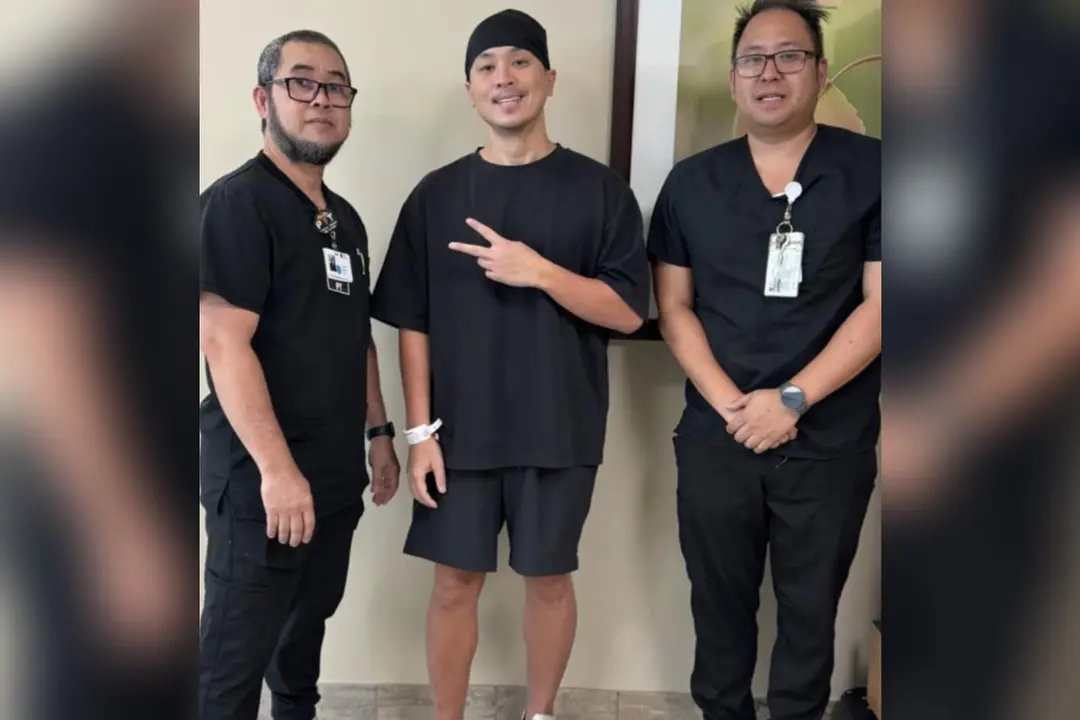 Patient in black makes peace sign while standing between two medical staff in black scrubs with ID badges.