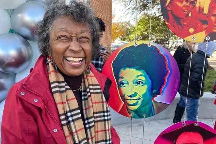 Smiling woman in red jacket and plaid scarf beside colorful circular portraits displayed at an outdoor event.