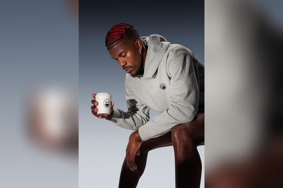 Man with red braids in grey hoodie holds a white coffee cup with a smiley-face logo.