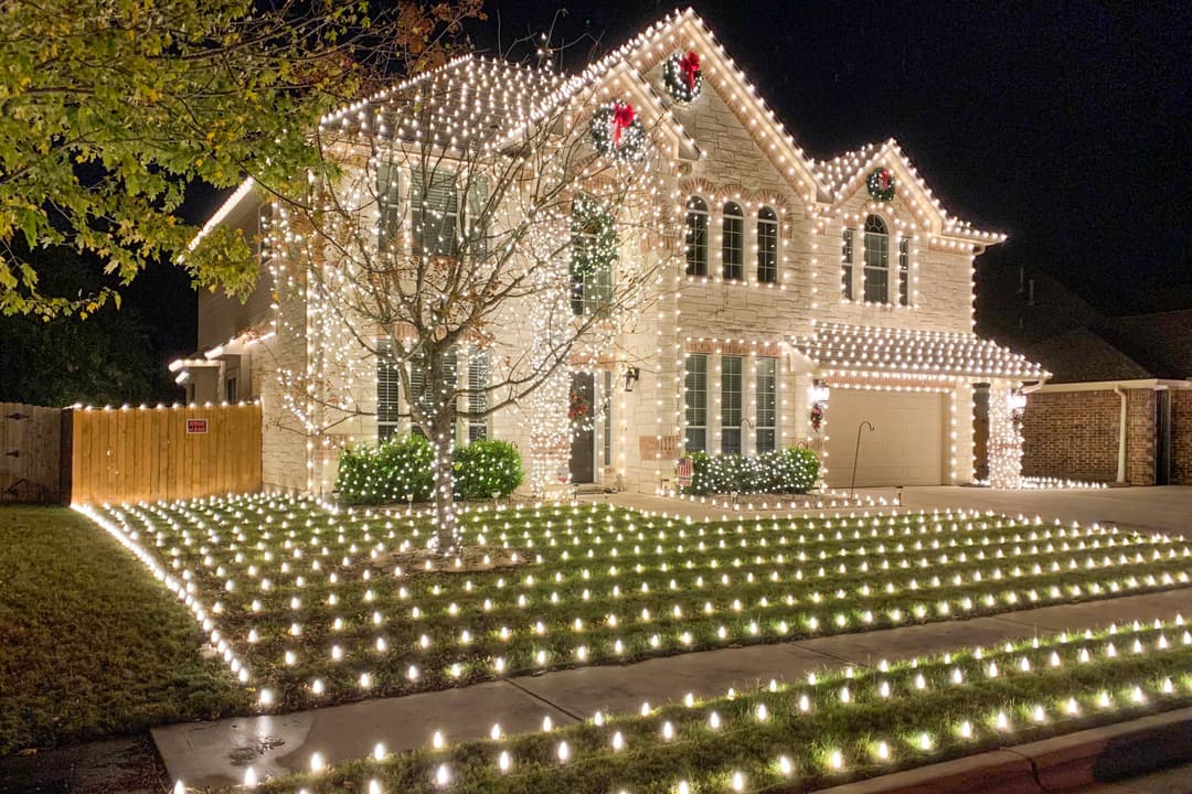 Two-story stone house covered in white Christmas lights at night, with wreaths and a lawn full of lights.