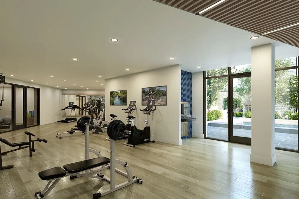 Modern fitness center with weight benches, exercise bikes, and treadmills beside large windows overlooking a courtyard.