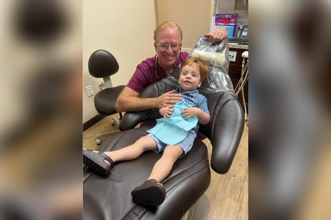 Smiling dentist poses beside a young boy seated in a dental chair wearing a blue bib.