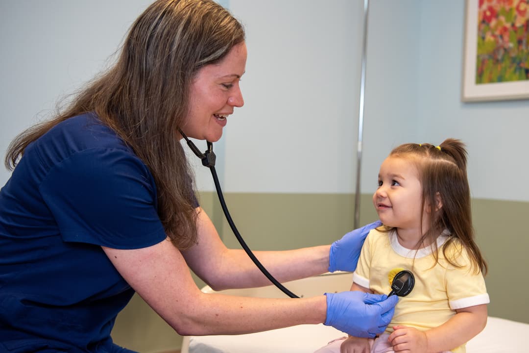 Smiling nurse in blue scrubs uses a stethoscope to listen to a young girl's heartbeat during a checkup.
