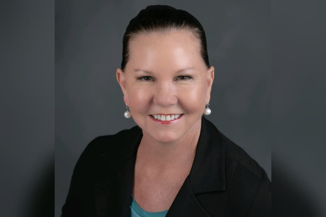Smiling woman with pulled-back hair, pearl earrings and black blazer against gray backdrop.