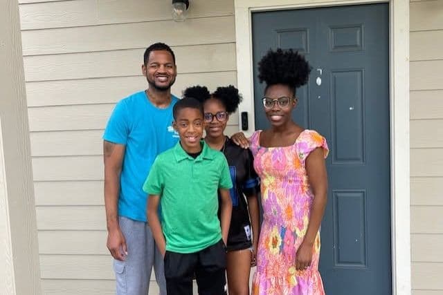 Family of four smiling outside a home's front door with a gray door and beige siding.