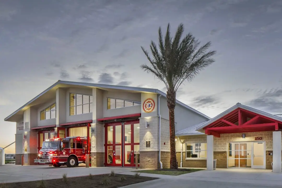 Modern fire station at dusk with a fire truck in an open bay, palm tree out front, and red-trimmed entrance.