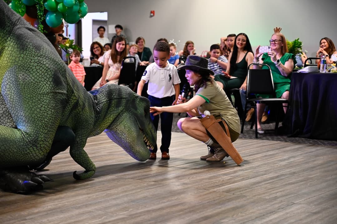 A boy and a costumed presenter with a microphone meet a large dinosaur puppet as an audience watches.