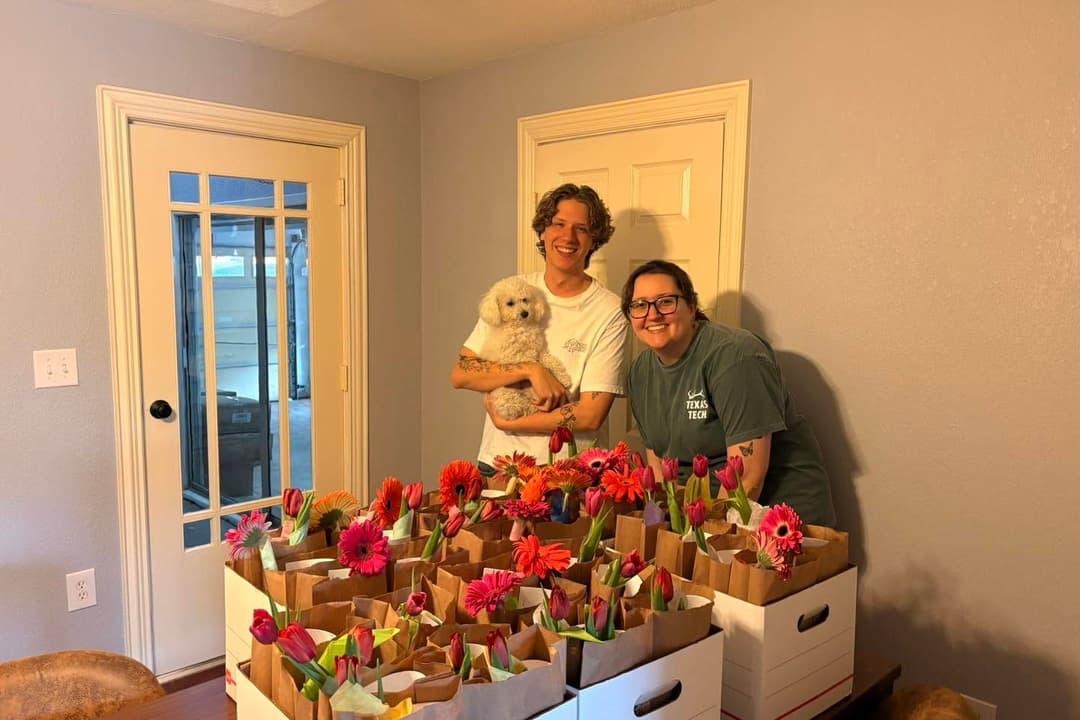 Smiling couple holding a white dog behind boxes of flower bouquets in paper bags indoors.