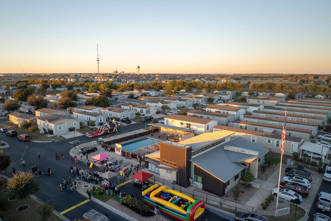Aerial view of community festival with bounce houses, tents, and pool outside a clubhouse in a mobile home park at sunset.