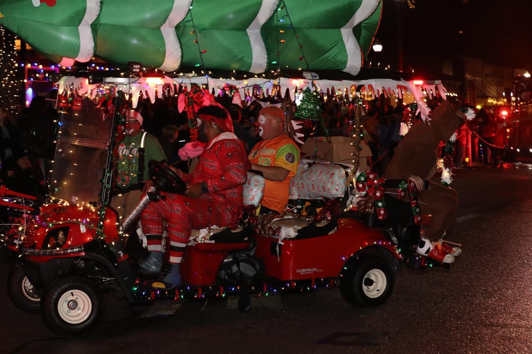 Clowns ride a festively lit golf cart decorated with lights and gifts in a nighttime Christmas parade.