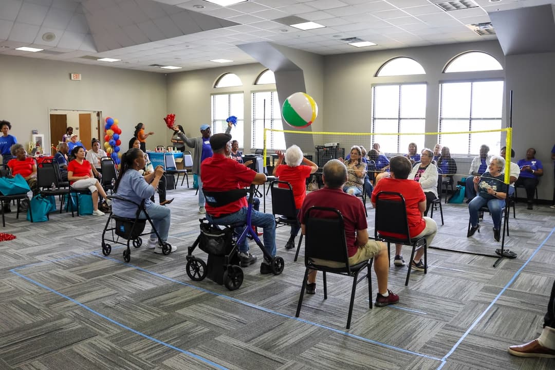 Seniors seated play beach ball volleyball over a net in a community room during a group activity.