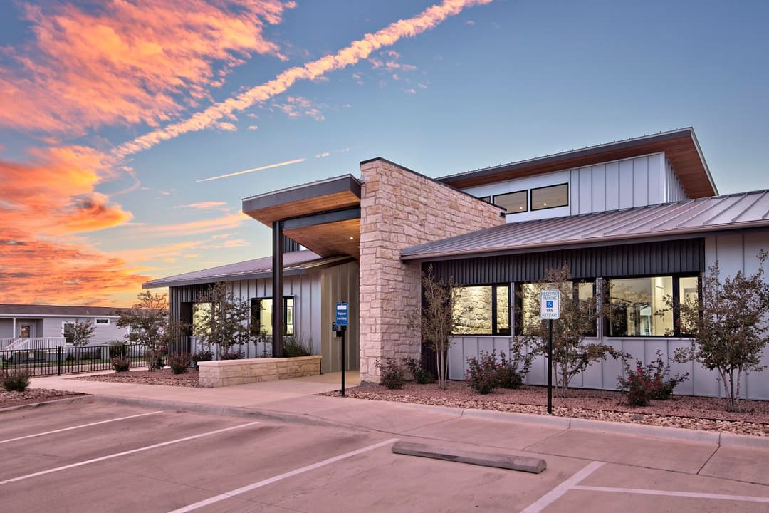 Modern single-story building with stone and metal siding, large windows, and parking lot at sunset.