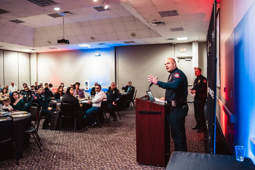 Uniformed officer speaks at a podium to a seated audience in a banquet hall during an event.