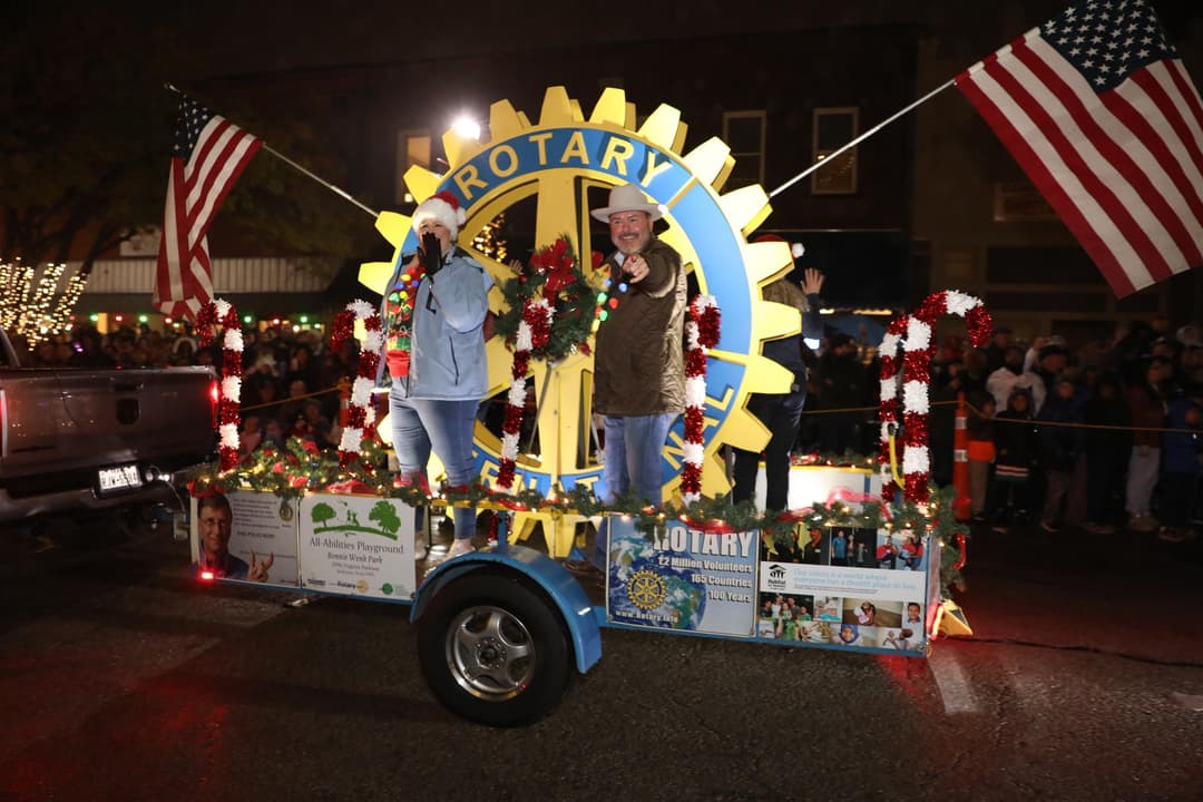 Rotary Club Christmas parade float with American flags, lit decorations, and two people waving at a nighttime crowd.