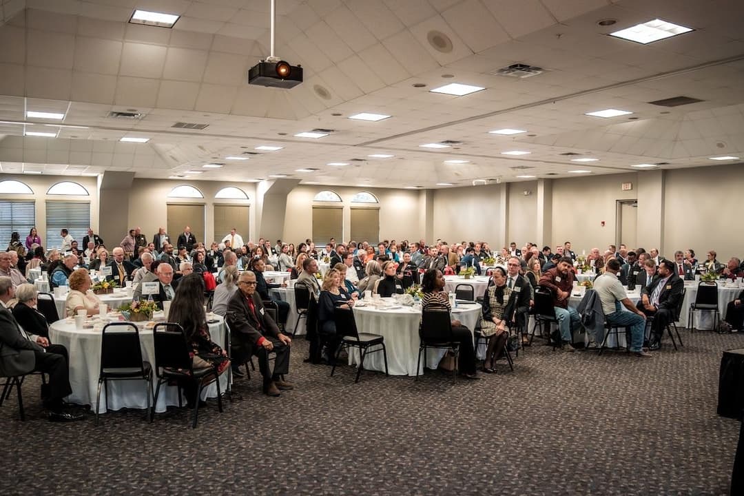 Large crowd seated at round tables with white cloths in a banquet hall during an event.