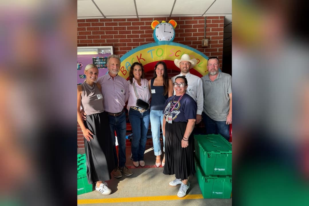 Seven adults pose by a "Back to School" inflatable arch and green bins at a school donation event.