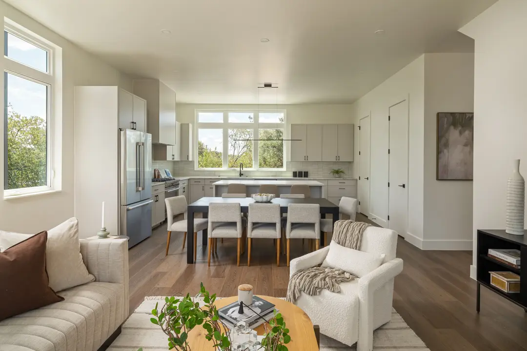 Open-concept living room, dining table with eight chairs, and modern kitchen with large windows and wood floors.