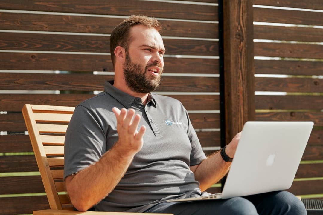 Bearded man in gray polo gesturing while using a laptop outdoors against a wooden slat wall.