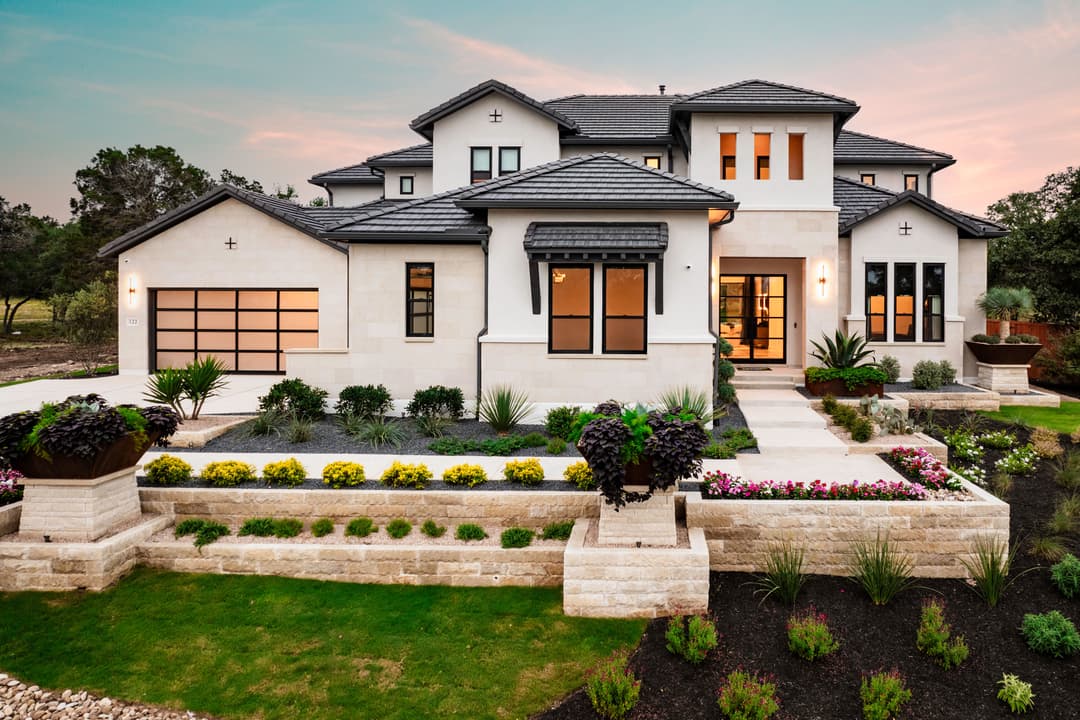 Luxury two-story stucco home with tile roof, lit windows at dusk, terraced stone landscaping and manicured gardens.