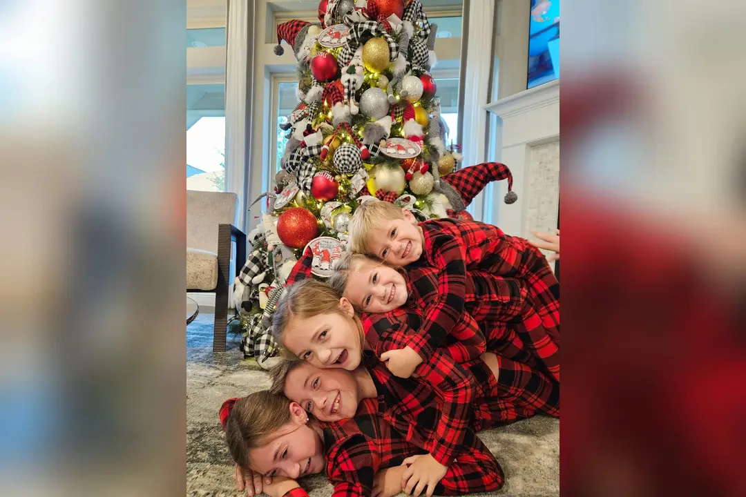 Five children in matching red plaid pajamas smile while piled together in front of a decorated Christmas tree.