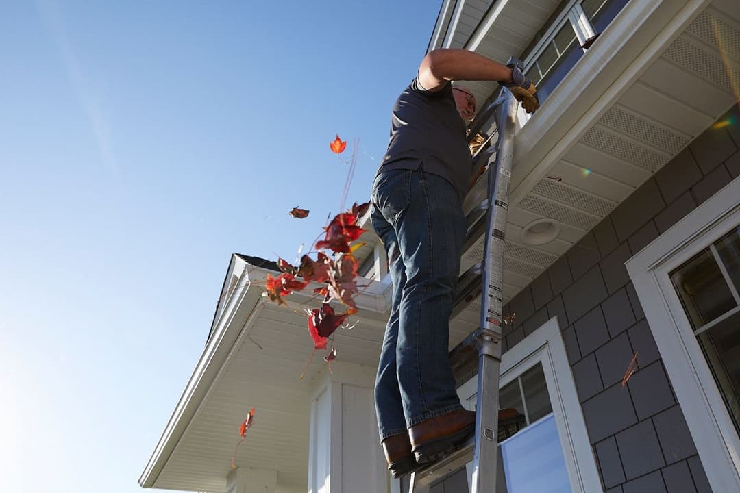 Man on a ladder cleaning leaves from a house gutter, tossing fallen red leaves to the ground.