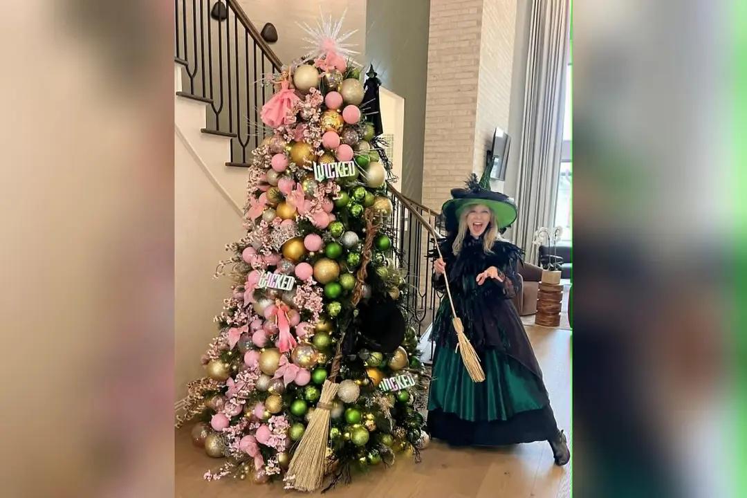 Woman in witch costume with broom beside Wicked-themed Christmas tree decorated in pink and green ornaments.