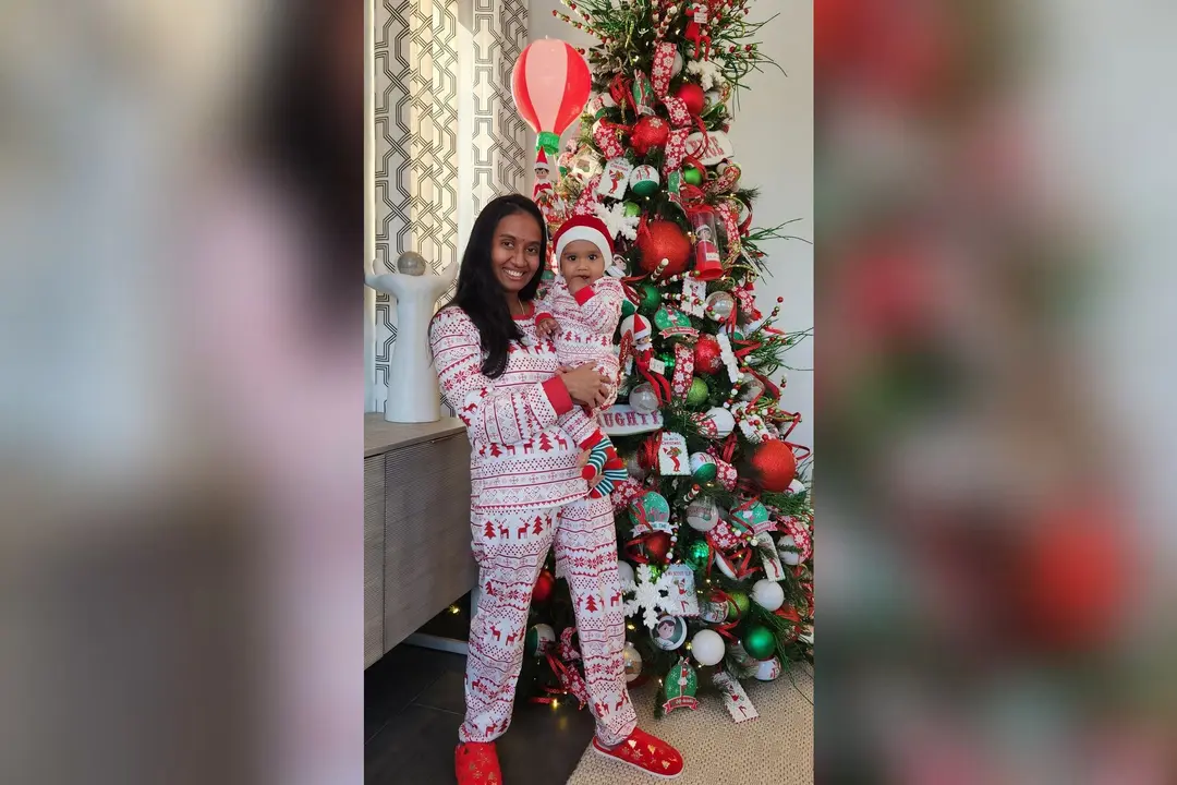 Smiling woman holding a baby in a Santa hat, both in matching pajamas, beside a decorated Christmas tree.