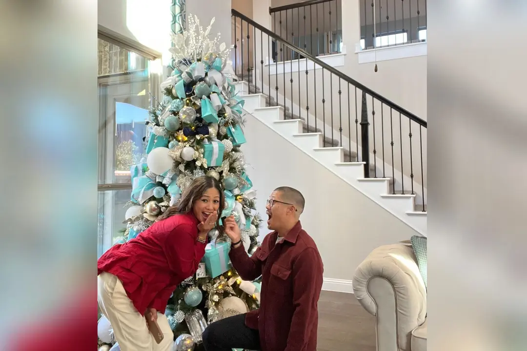 Couple reacting with surprise beside a teal and white decorated Christmas tree near a staircase.