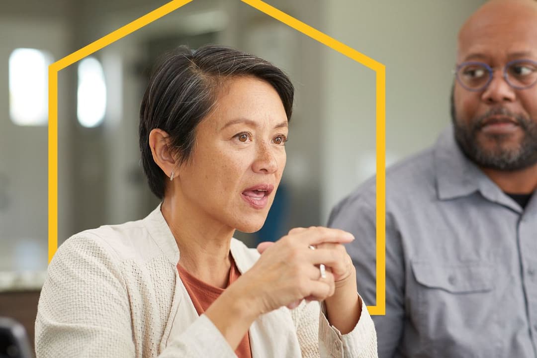 Woman gestures while speaking, man in glasses listening beside her, with yellow house-shaped outline overlay.