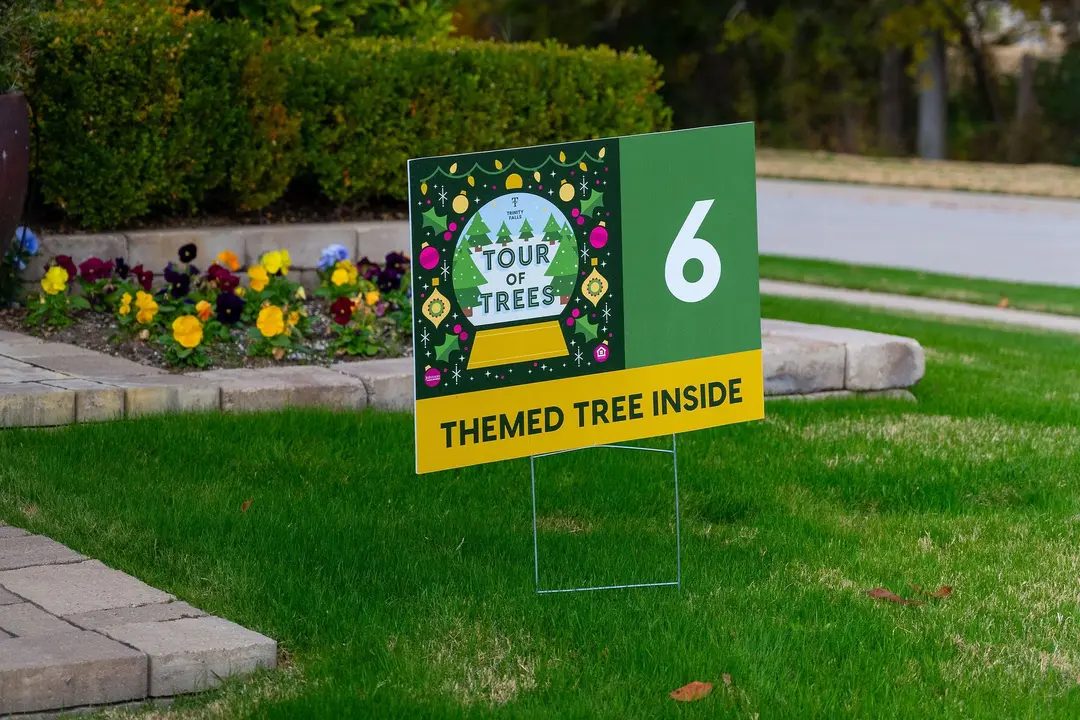 Yard sign reading "Tour of Trees 6, Themed Tree Inside" on a lawn near a flower bed.