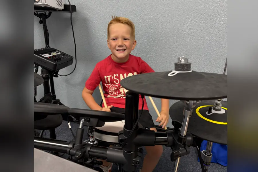 Smiling young boy in a red shirt plays an electronic drum set, holding drumsticks.