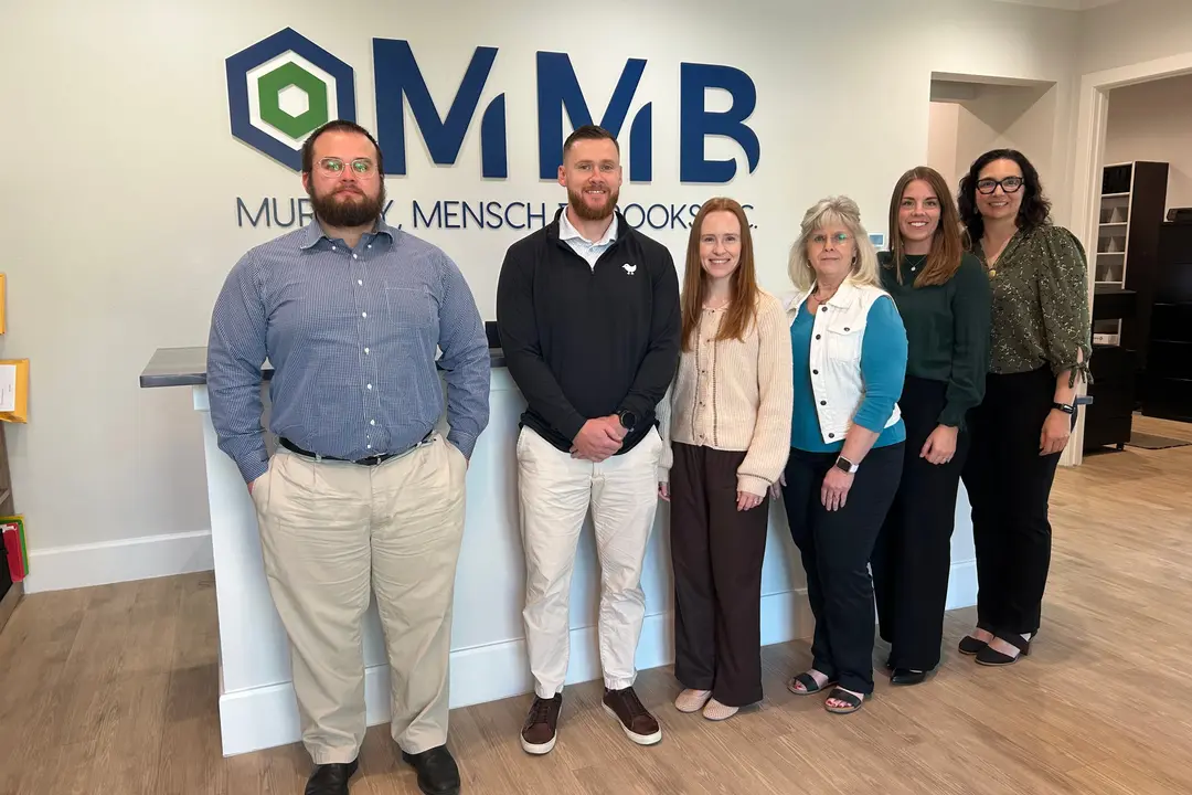 Six people stand together in front of an MMB (Murphy, Mensch & Brooks) office wall logo.