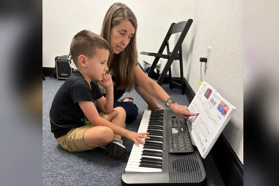 A woman teaches a young boy to play a keyboard while pointing at sheet music during a piano lesson.