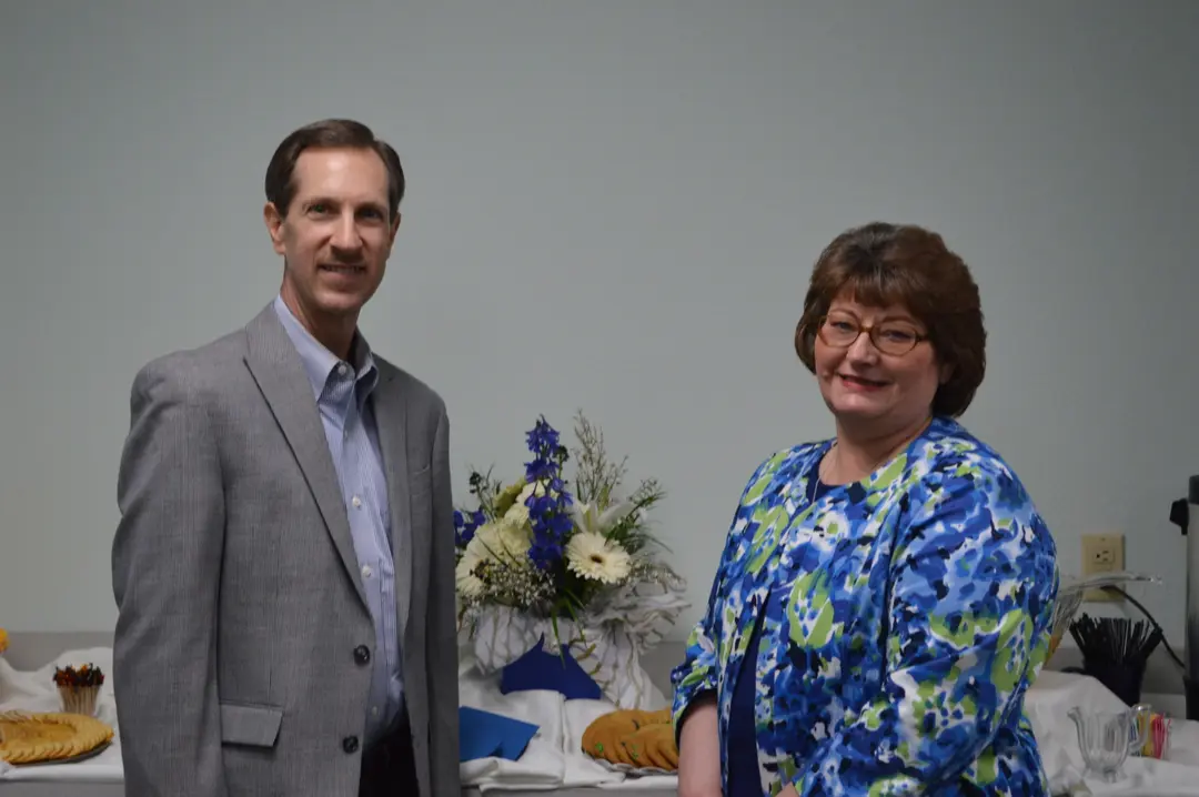 A man in a gray blazer and a woman in a blue floral top stand beside a table with flowers and cookies.