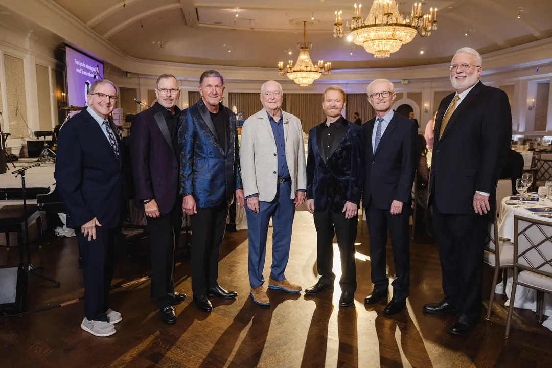 Seven men in suits pose together at a formal ballroom event with chandeliers and set dining tables.